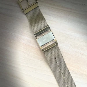 Guldfärgad klocka från DKNY - Snygg guldfärgad klocka från DKNY med ett stilrent mesh-armband. Klockan har en rektangulär urtavla och ett justerbart spänne. Perfekt för att ge en elegant touch till din outfit.