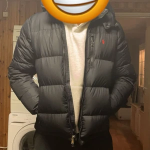 Svart pufferjacka från Ralph Lauren - Säljer en svart pufferjacka från Ralph Lauren med dragkedja och huva. Jackan har en broderad logga på bröstet och är perfekt för kyligare väder. Den har långa ärmar och en stilren design.