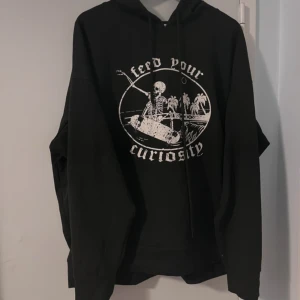 Svart hoodie med skelettmotiv - Snygg svart hoodie med ett coolt skelettmotiv och texten 'Feed Your Curiosity'. Perfekt för en avslappnad stil med en touch av edge. Hoodien har långa ärmar och en klassisk huva med dragsko. Väldigt lätt material, är ganska gammal men har inga skador. 