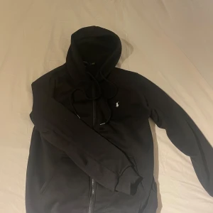 Svart hoodie från Polo Ralph Lauren - Säljer en svart hoodie från Polo Ralph Lauren med dragkedja och en liten broderad logga på bröstet. Tröjan har en avslappnad passform och är perfekt för en casual look. Den har långa ärmar och en justerbar huva med snören.