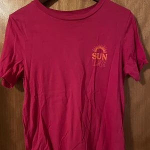 Rosa t-shirt från Gina Tricot - Säljer en rosa t-shirt från Gina Tricot med trycket 'SUN DAY' i orange på bröstet. T-shirten har en klassisk rund hals och korta ärmar. Perfekt för en avslappnad stil.