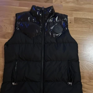 Svart dunväst från Moncler - Snygg svart dunväst från Moncler med glansig yta och dragkedja framtill. Västen har två praktiska fickor med dragkedjor och en diskret logga på bröstet. Perfekt för kyligare dagar.