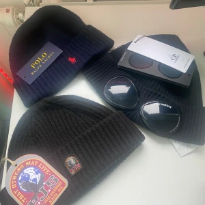 Svarta mössor från Polo Ralph Lauren, CP Company och PJS - Säljer tre svarta ribbade mössor från Polo Ralph Lauren, CP Company och PJS. Mössorna är stilrena och passar perfekt för kyligare väder. De har en klassisk design med uppvikt kant och är tillverkade i mjukt material för extra komfort.