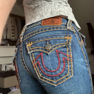 Lågmidjade true religion jeans - Snygga blå true religion jeans med unika broderade fickor i rött. De har en låg midja och en bootcut passform. Perfekta för en avslappnad stil med coola detaljer som knappar och kontrastsömmar. De är i storlek 12 och skulle säga att de passar någon som ofta när xs/S, 32 eller 25. Kom gärna med prisförslag!