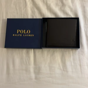 Brun plånbok från Polo Ralph Lauren - Snygg brun plånbok från Polo Ralph Lauren i skinn. Den har flera kortfack och en myntficka med knapp. Kommer i en elegant blå förpackning med guldfärgad text.