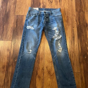 Blå jeans med slitningar från Hollister - Säljer ett par snygga blå jeans från Hollister med coola slitningar på framsidan. De har en knappgylf och klassisk femficksdesign. Perfekta för en avslappnad stil.