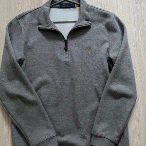 Grå tröja med dragkedja från Polo Ralph Lauren - Säljer en stilren grå tröja från Polo Ralph Lauren med en liten broderad logotyp på bröstet. Tröjan har en praktisk dragkedja vid halsen och långa ärmar. Perfekt för en avslappnad och snygg look.
