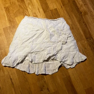 Vit omlottkjol med volang - Charmig vit omlottkjol med volangdetaljer. Perfekt för en somrig look. Kjolen har en luftig och lätt design som ger en avslappnad känsla.