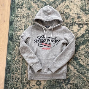 Grå hoodie från superdry. - Säljer en snygg grå hoodie från Superdry med broderad text i marinblått och rött på framsidan. Tröjan har en klassisk känguruficka och justerbar huva med vita snören. Nypris kostar den ungefär 1500 kronor nu lägger jag ut den för 500kr. Perfekt för en avslappnad stil.