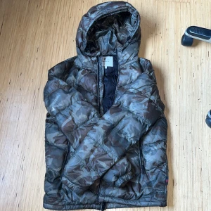  Woolrich Camouflage pufferjacka  - Säljer en woolrich camouflage pufferjacka med huva. Jackan har en dragkedja framtill och är perfekt för kyligare dagar. Den har ett klassiskt camo-mönster i gröna och bruna toner. Jackan har två små hål. Ett bakom nertill och ett på H. Arm 