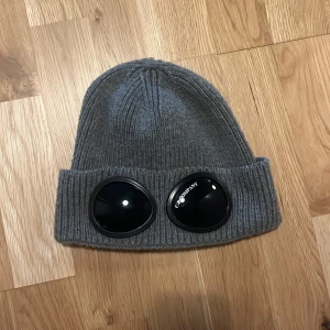 Grå mössa från C.P. Company - Snygg grå mössa från C.P. Company med unika glasögondetaljer på uppviket. Tillverkad i mjuk bomull och perfekt för kyliga dagar. Pris ej hugget i sten 