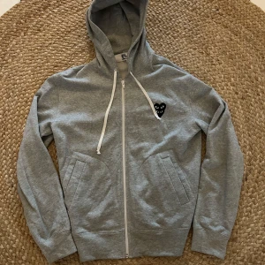 Grå hoodie från Comme des Garçons - Säljer en stilren grå hoodie från Comme des Garçons med dragkedja och det ikoniska hjärta-emblemet på bröstet. Tröjan har en justerbar huva med vita snören och praktiska fickor framtill. 