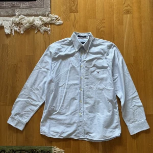 Randig skjorta från GANT - Snygg randig skjorta från GANT i ljusblått och vitt. Skjortan har en klassisk krage och knappar framtill. Den är långärmad och har en broderad logga på bröstfickan. Perfekt för en stilren look!
