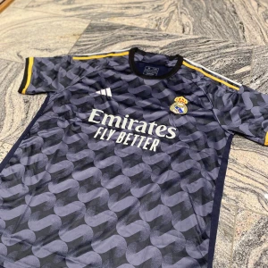 Mönstrad fotbollströja från Adidas - Snygg fotbollströja från Adidas med Real Madrid-logga. Tröjan är mörk med ett unikt mönster och har korta ärmar med gula detaljer. Perfekt för fans av laget! Storlek M!