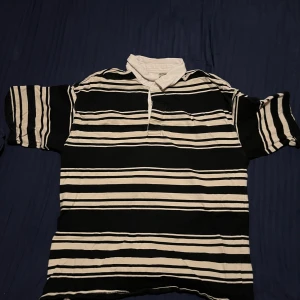 Cream och svart Polo shirt  - Säljer en klassisk svart och cream randig pikétröja med korta ärmar. Perfekt för en avslappnad stil. Tröjan har en krage och knappar framtill. Passar bra till jeans eller shorts.