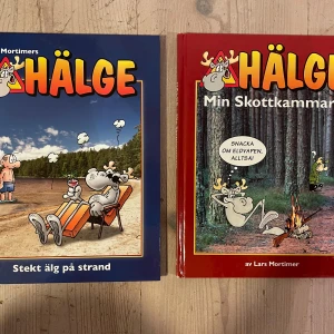 Hälge - Stekt älg på strand och Min Skottkammare - Två humoristiska serieböcker av Lars Mortimer med älgen Hälge i huvudrollen. 'Stekt älg på strand' har ett blått omslag och 'Min Skottkammare' har ett rött omslag. Perfekt för den som gillar skämt och serier om skogen och jakt. Mycket fina välbehållna nästintill oanvända inbundna böcker.
