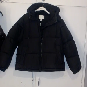 Svart dunjacka från H&M - Säljer en svart dunjacka från H&M, helt ny med prislapp. Med dragkedja och huva. Jackan är perfekt för kalla dagar och har en pufferdesign som håller dig varm. Den har en klassisk look som passar till det mesta. Perfekt för vintern! ❤️