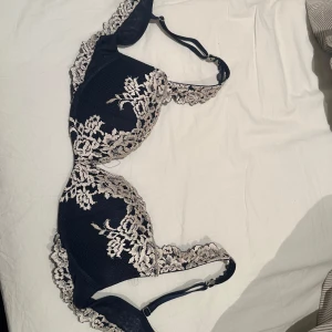 Blå spets-bh från Intimissimi - Säljer en helt oanvänd bh. Det är elegant blå bh från Intimissimi med vacker vaniljfärgad spetsdetalj. Bh:n har justerbara axelband och en bekväm passform. Perfekt för att känna sig extra fin under kläderna. Säljer denna bh för att den är en storlek för stor för mig.