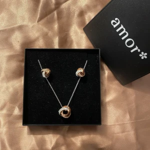 Smyckeset från Amor - Elegant smyckeset från Amor med ett halsband och matchande örhängen. Designen har en tvinnad look i silver och guld, vilket ger en stilren och modern känsla. Perfekt för att lyfta vilken outfit som helst.