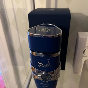 Asad Zanzibar Eau de Parfum från Lattafa - Helt ny bara använt ungefär 5 spray. Pris kan diskuteras. Elegant parfymflaska i djupblått med silverdetaljer och ett dekorativt mönster. Asad Zanzibar från Lattafa erbjuder en sofistikerad doftupplevelse. Flaskan har en unik design med silverband och ett intrikat emblem på framsidan.