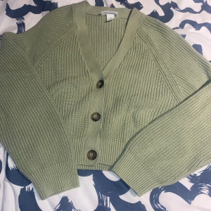 Grön ribbad kofta från Monki - Snygg grön kofta från Monki med ribbad design och stora knappar framtill. Den har en croppad passform och långa ärmar, perfekt för lager på lager-stilen. Ett stilrent plagg som funkar till många olika outfits.