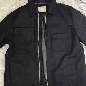 Svart Overshirt/Jacka från Jack & Jones Premium - Säljer nu denna snygga Overshirt från Jack & Jones Premium, endast prövad och aldrig andvänd ute i sol eller regn. säljer den då det var ett spontanköp och jag inte riktigt tyckte det va nått för mig, finns inga defekter eller skador överhuvudtaget. Nypris är runt 1.200kr men säljer den för 900kr