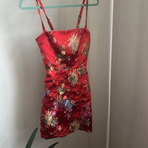 Röd blommig klänning med justerbara axelband - Snygg röd klänning i satin med blommigt mönster och justerbara axelband. Perfekt för en färgglad look med vackra detaljer i guld och andra färger. Klänningen har en smickrande passform och är kort i modellen. Storlek XS från urban outfitter, nypris 450kr 