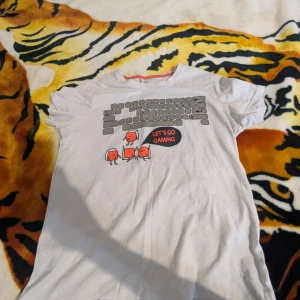 Vit t-shirt med gaming-tryck från Lindex - Cool vit t-shirt från Lindex med ett roligt gaming-tryck som säger 'Let's Go Gaming'. Perfekt för unga gamers som vill ha en bekväm och stilren look. Tillverkad i ett hållbart materialval.