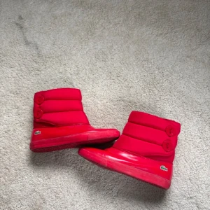 Vintage Röda boots från Lacoste Stl 40 - Snygga röda boots från Lacoste med quiltad design och rund tå. De har en vadderad look och är perfekta för kyligare dagar. Skorna har en diskret Lacoste-logga på sidan och en robust sula för bra grepp.