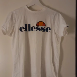 Vit t-shirt från Ellesse - Säljer en klassisk vit t-shirt från Ellesse med deras ikoniska logga i blått och orange på bröstet. T-shirten har en rund hals och korta ärmar, perfekt för en avslappnad stil.