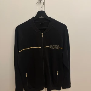 kofta Hugo Boss - Snygg svart kofta från Hugo Boss med dragkedja och gulddetaljer. Jackan har långa ärmar och två fickor med dragkedjor. 