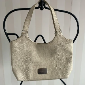 Beige axelväska från Betty Barclay - Snygg beige axelväska från Betty Barclay med krokodilpräglat mönster. Väskan har dubbla handtag och en praktisk dragkedja upptill. Perfekt för att ge en stilren touch till din outfit.