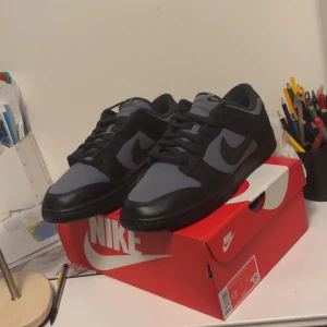 Nike Dunk i svart och grått - Snygga Nike Dunk sneakers i en stilren kombination av svart och grått. Skorna har en klassisk design med snörning och en robust sula. Perfekta för en casual look.