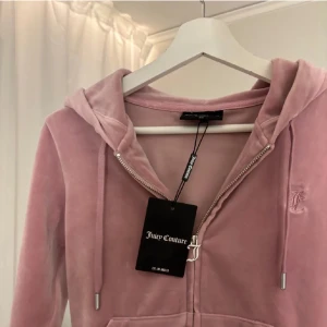 Rosa och svart hoodie från Juicy Couture - Säljer en snygg rosa hoodie i sammet från Juicy Couture med dragkedja och huva. Den har en broderad logga på bröstet. Baksidan är svart med glittrig Juicy Couture-text. Perfekt för en avslappnad stil.