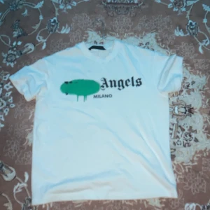 Vit t-shirt från Palm Angels - Säljer en vit t-shirt från Palm Angels med ett grönt grafiskt tryck och texten 'Milano' på framsidan. T-shirten har en klassisk rund hals och korta ärmar. Perfekt för en avslappnad stil.