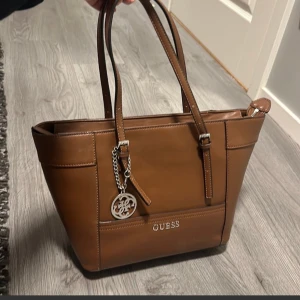 Guess handväska - Bra skick se bild för att avgöra. Har använt den några få gånger sen har den bara stått i garderoben  GUESS DELANEY SMALL CLASSIC TOTE Storlek: ONE SIZE Färg: COGNAC  Length 29 cm Height 24.5 cm Depth 15 cm