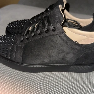 Louboutin skor  - Snygga svarta sneakers från Christian Louboutin i mocka med nitar på tån och den ikoniska röda sulan. Skorna har snörning och en stilren design som sticker ut. Lite mer använda än på bilden. 