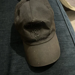 Svart keps från Moschino - Snygg svart keps från Moschino med broderad logga framtill. Kepsen har en justerbar rem baktill för perfekt passform. Perfekt för en stilren look.