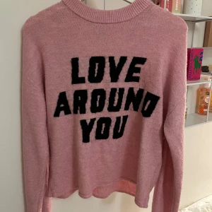 Rosa stickad tröja med text - Säljer en rosa stickad tröja med texten 'LOVE AROUND YOU' i svart på framsidan. Tröjan har långa ärmar och en rund halsringning. Perfekt för en mysig och stilren look.