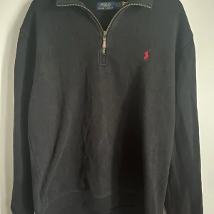 Svart tröja med dragkedja från Polo Ralph Lauren - Säljer en stilren svart tröja från Polo Ralph Lauren med en halv dragkedja och det klassiska röda broderade logotypen på bröstet. Perfekt för en avslappnad men ändå snygg look. Tröjan har långa ärmar och är gjord i ett bekvämt material.