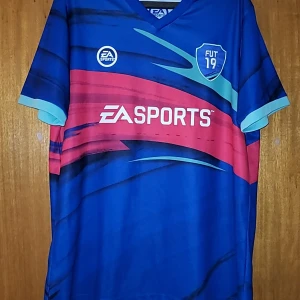 Blå fotbollströja från EA Sports FIFA 19 - Snygg blå fotbollströja från EA Sports med korta ärmar och ett coolt mönster i rött och turkost. Tröjan har en V-ringning och ett stort nummer 19 på ryggen. Perfekt för gamingfans och fotbollsälskare!