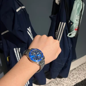 Blå och silverfärgad klocka - Snygg klocka med blå urtavla och silverfärgad metallarmband. Klockan har en blå bezel med vita markeringar och romerska siffror på urtavlan. Perfekt accessoar för att ge en stilren touch till din outfit.