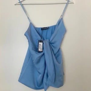 Blå satintopp från prettylittlething  - Säljer en ljusblå satin topp från prettylittlething. Toppen har smala axelband och en knytdetalj framtill.  Prislapp kvar