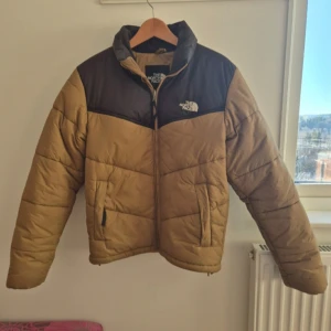 Beige och svart dunjacka från The North Face - Säljer en snygg beige och svart dunjacka från The North Face. Jackan har en klassisk pufferdesign med dragkedja och hög krage för extra värme. Perfekt för kyliga dagar. 🧥