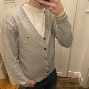 Grå kofta från Tommy Hilfiger - Säljer en stilren grå kofta från Tommy Hilfiger i premium bomull. Koftan har knappar framtill och en liten logga på bröstet. Passar till både jeans och chinos. Modellen är ca 182, tveka inte att kontakta vid eventuella frågor 😀