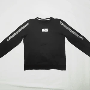 Svart sweatshirt från Emporio Armani - Säljer en stilren svart sweatshirt från Emporio Armani i storlek 150cm med deras logga på bröstet och längs armarna. Tröjan är sparsamt använd och jag skulle säga att den är i skick 8/10. Etiketten i nacken har dock trillat av men kvitto medföljer. Tröjan är köpt i butik, ”Room 1006” - Malmö och är 100% äkta. Nypris 699kr, Jan säljer den för endast 349kr! Original förpackning medföljer! Skriv om du har fler frågor!