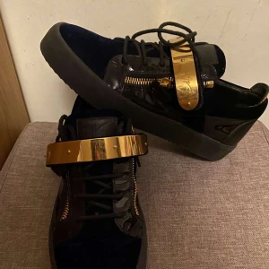  zanotis skor  - Snygga svarta sneakers med glänsande gulddetaljer och snörning. Skorna har en unik design med guldfärgad rem över vristen och dragkedjor på sidorna. Perfekta för att ge en lyxig touch till din outfit.