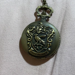 Fickur med Ravenclaw-emblem - Snyggt fickur med ett detaljerat Ravenclaw-emblem på framsidan. Uret har en antik finish med en kedja för att enkelt bära runt halsen. Insidan visar en klassisk urtavla med svarta siffror och dekorativa visare. Klockan är trasig men borde inte vara svår att laga, har en liten färgfläck på baksidan.