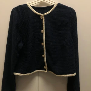 Mörkblå kavaj med kjol - Elegant mörkblå kavaj med matchande kjol. Kavajen har långa ärmar och knäpps med stilrena knappar framtill. Både kavajen och kjolen har dekorativa guldfärgade kanter som ger en lyxig touch. Perfekt för en stilren look.