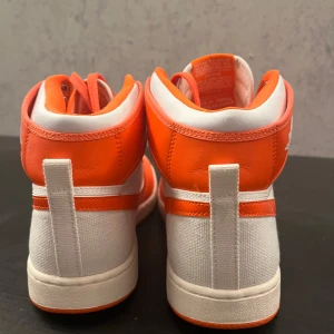Nike sneakers i vitt och orange - Snygga Nike sneakers i en fräsch kombination av vitt och orange. Skorna har en klassisk design med snörning och den ikoniska Nike-loggan på sidan. Perfekta för en sportig look.